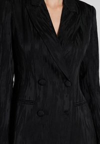 Blazer negro de doble botonadura con una tela texturizada con patrones de olas verticales, solapas muescas y dos botones grandes en el frente.