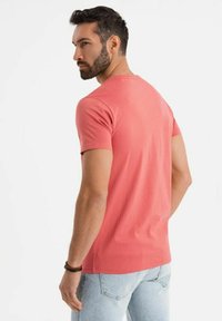 Tričko s krátkým rukávem a kulatým výstřihem v korálové barvě, vyrobené z bavlny, s hladkou texturou a přiléhavým střihem. Nosené s světle modrými džíny.