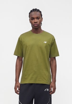Man met gevlochten haar draagt een olijfgroen New Balance T-shirt en zwarte sportshorts met logo's, staand tegen een effen witte achtergrond.
