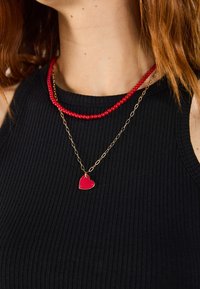 Collar de cuentas rojas y cadena dorada con un pequeño colgante en forma de corazón rojo, superpuesto a una blusa negra con textura acanalada. Textura suave y acabado brillante.