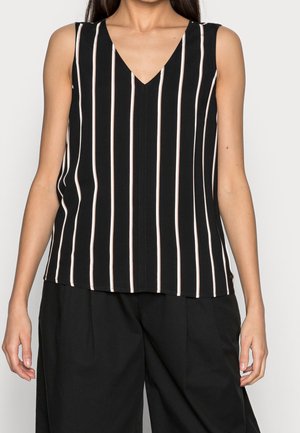 Vrouw draagt een mouwloze zwarte top met witte verticale strepen en zwarte broek met wijde pijpen tegen een effen achtergrond.