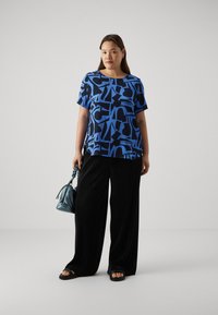 Blue short-sleeve top with abstract black patterns, paired with black wide-leg pants. Complemented by a teal handbag and black sandals.