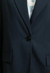 Marinblå kavaj med en mjuk textur, enkel svart knapp och två framfickor. Har en subtil lyster och en skräddarsydd passform.