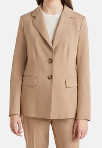 Béžový blazer se dvěma předními knoflíky, s výstřihem do V a dvěma předními kapsami, vyrobený z hladké, lehké tkaniny. Stylizovaný s lehkou košilí pod ním.