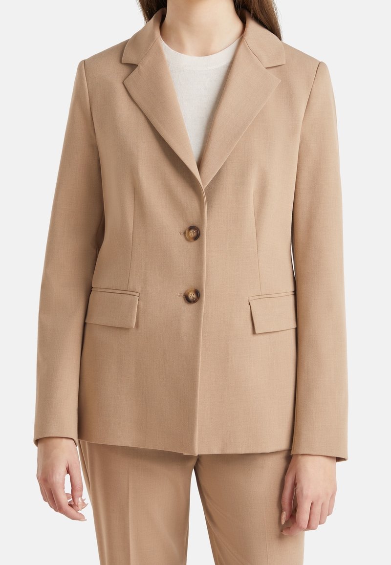 Béžový blazer se dvěma předními knoflíky, s výstřihem do V a dvěma předními kapsami, vyrobený z hladké, lehké tkaniny. Stylizovaný s lehkou košilí pod ním.