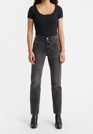 Femme portant un haut noir côtelé à manches courtes, un jean noir délavé taille haute et des bottines noires, debout devant un fond uni.