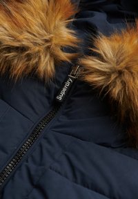 Veste matelassée bleu marine avec une fermeture éclair frontale et une bordure en fausse fourrure marron autour de la capuche, tirette de fermeture éclair indiquant "Superdry."