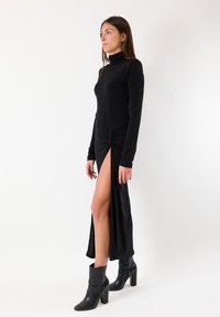 Abito nero a maniche lunghe con collo alto, caratterizzato da uno spacco alla coscia e dettaglio arricciato in vita. Indossato con stivaletti neri.