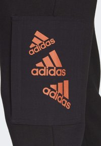 Černé tepláky s velkou obdélníkovou oranžovou kapsou, na které jsou zobrazeny tři naskládané loga Adidas. Hladká tkanina s prošívanými detaily.