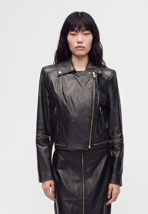 Femme aux longs cheveux noirs portant une veste en cuir noire avec des fermetures éclair dorées et une jupe en cuir noire assortie sur un fond uni.