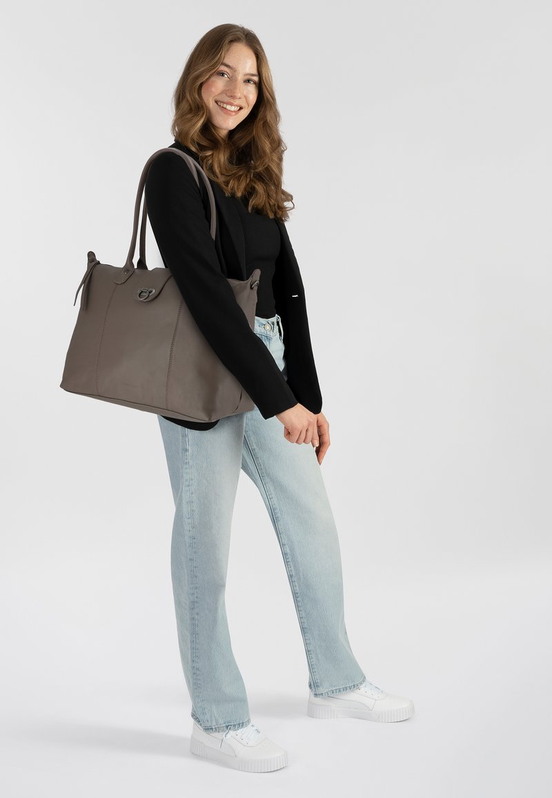 Donna sorridente che indossa un top nero, jeans azzurri chiari, sneakers bianche e giacca nera, mentre porta una grande borsa a tracolla grigia su uno sfondo bianco.