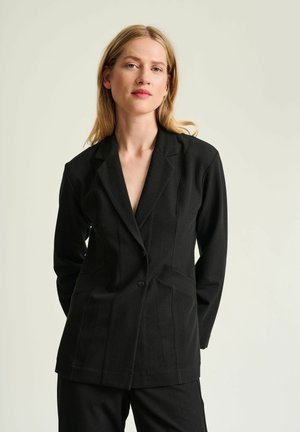Mujer con cabello rubio que lleva una blazer negra ajustada, de pie frente a un fondo claro y liso, mirando a la cámara.