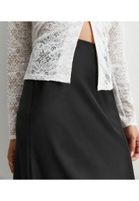 Witte kanten top met lange mouwen, gecombineerd met een zwarte satijnen rok. De kant heeft bloemmotieven en delicate randen; de rok is soepel en elegant.