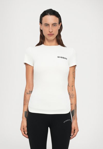 Femme aux longs cheveux noirs, portant un t-shirt blanc ajusté à manches courtes et des leggings noirs avec des tatouages visibles sur les deux bras, faisant face à l'avant.
