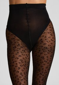 Schwarz-hochgeschnittene Strumpfhosen mit Leopardenmuster von der Mitte des Oberschenkels bis zu den Knöcheln, getragen von einer stehenden Person vor einem schlichten Hintergrund.