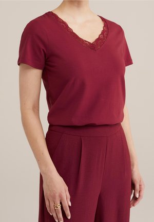 Femme portant un haut bordeaux à manches courtes avec un col en V en dentelle et un pantalon plissé taille haute assorti, montrant le côté gauche et une main avec une bague.