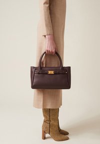 Braune Lederhandtasche mit zwei Griffen, goldenen Beschlägen und strukturiertem Finish, gehalten von einer Person in einem hellbeigen Kleid und gemusterten knielangen Stiefeln.
