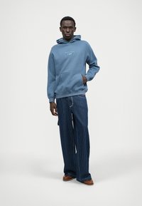 Felpa blu con tasca frontale e cordini, abbinata a pantaloni larghi a righe blu. Le scarpe marroni completano il look.