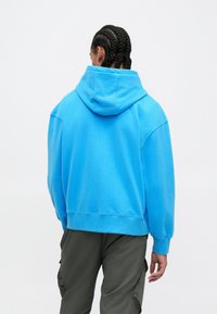 Osoba s pletenom kosom, odjevena u svijetloplavi hoodie i tamne cargo hlače, stoji okrenuta leđima prema jednostavnoj bijeloj pozadini.