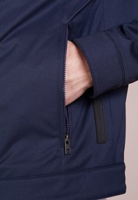 Marineblå jakke i blødt, struktureret materiale har en lynlåslomme og en sort læderlabel med "Michael Kors" på siden.