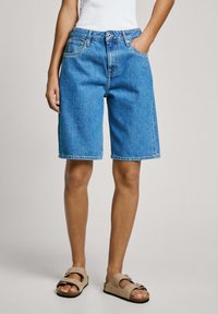Mujer con pantalones cortos de mezclilla azul hasta la rodilla, blusa blanca y sandalias beige con hebilla, de pie contra un fondo liso.