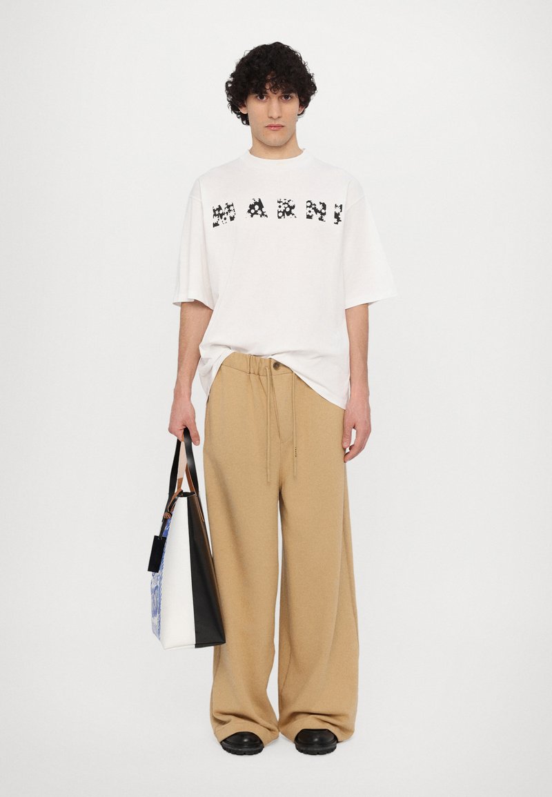 Weiße oversized T-Shirt mit schwarzem Grafiktext, kombiniert mit lockeren beigefarbenen Hosen. Model hält eine mehrfarbige Tragetasche mit schwarzen Akzenten.