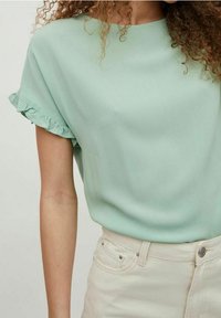 Blouse vert menthe avec manches courtes à volants, en tissu doux, associée à un pantalon de couleur claire. Le tissu a une texture lisse.