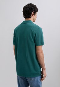 Polo shirt verde oscuro de algodón, de manga corta, con cuello y dobladillo recto. Llevado con jeans azules, mostrando un diseño casual.