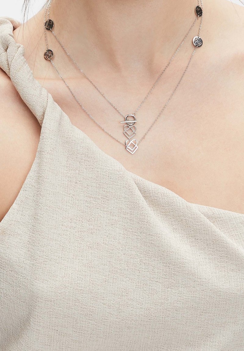 Deux colliers pendentifs superposés en forme de cœur argenté portés avec un haut beige texturé à une épaule, laissant apparaître la zone de la clavicule.