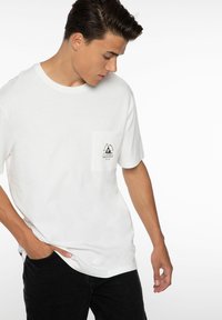 Witte katoenen t-shirt met ronde hals en korte mouwen. Heeft een linker borstzak met een zwart bergafbeelding logo.