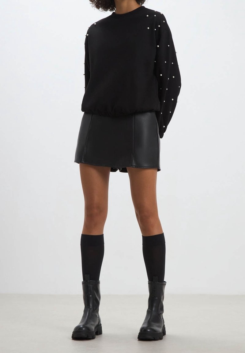 Femme portant un pull noir avec des détails en perles, une mini-jupe en cuir noir, des chaussettes montantes et des bottines noires, debout sur un sol gris.
