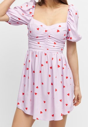 Femme portant une robe violet clair avec des manches bouffantes et des motifs de cœurs rouges, avec des détails froncés sur le corsage.