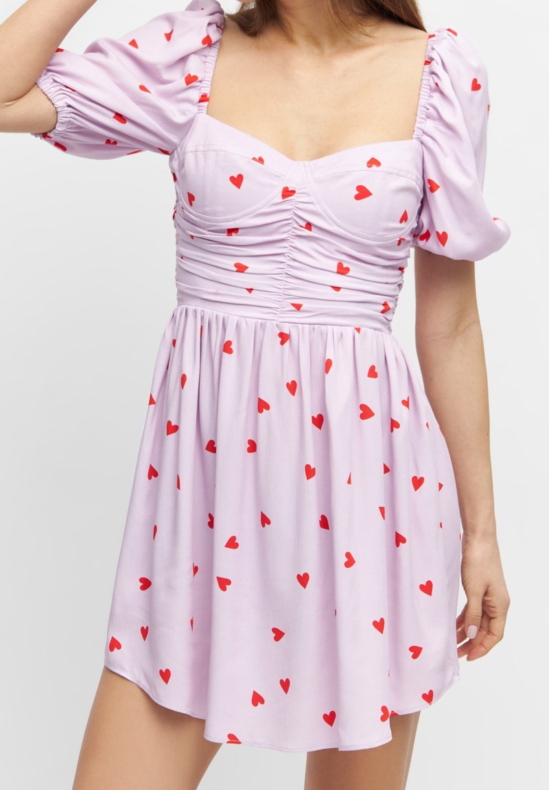 Femme portant une robe violet clair avec des manches bouffantes et des motifs de cœurs rouges, avec des détails froncés sur le corsage.