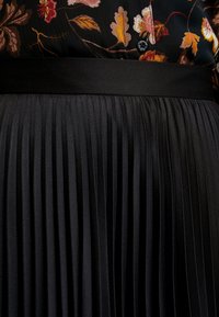 Svart plisserad kjol med en slät satinmidja, med vertikala plisseringar som skapar textur och rörelse.