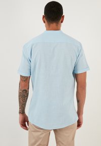 Camisa de manga corta en azul claro que presenta una tela texturada, puños doblados y un dobladillo curvado. El diseño es casual y transpirable.