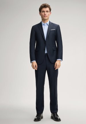 Młody mężczyzna stoi, ubrany w ciemnogranatowy garnitur z jasnoniebieską koszulą i czarnymi eleganckimi butami na jednolitym szarym tle.