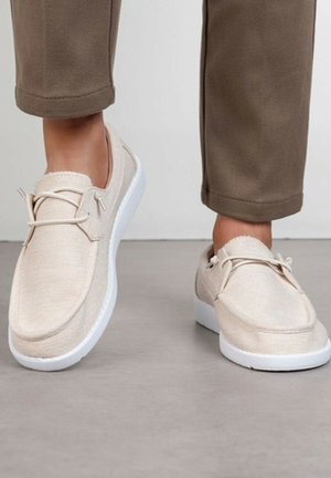 Pieds chaussés de chaussures beige à enfiler avec semelles blanches, posés sur un sol gris, assortis à un pantalon marron coupé.