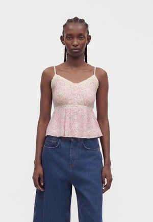 Kvinde med flettet hår iført en ærmeløs lyserød blomstret top med blondekant og løstsiddende mørkeblå jeans, der står mod en ensfarvet baggrund.