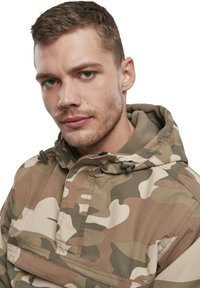Veste de camouflage avec des tons terreux de vert, marron et beige, dotée d'une capuche, d'une fermeture éclair et de détails de poches avant.