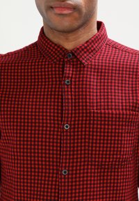 Camicia di flanella a quadri rossi e neri con colletto a pulsante, tasca anteriore e bottoni neri. Il tessuto ha una texture morbida.