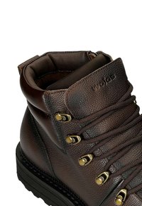 Hnědá kožená bota s texturovaným povrchem, kovovými očky a tkaničkami. Disponuje polstrovaným límcem a pevnou černou podrážkou.
