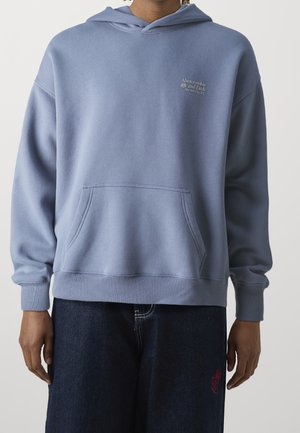 Osoba ubrana w jasnoniebieską bluzę z kapturem Abercrombie & Fitch z przednią kieszenią oraz ciemnoniebieskie dżinsy z czerwonym haftem na lewej stronie uda.