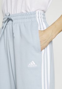 Pantalons de sport bleu clair avec une taille élastique, comportant le logo blanc d'Adidas et trois bandes blanches sur les côtés. Texture douce.