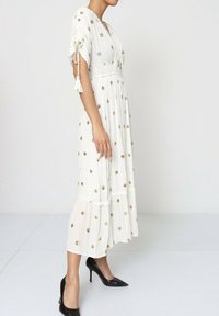 Robe maxi blanche à manches courtes, broderie florale dorée, design à volants, nouée à la taille, associée à des escarpins noirs à bout pointu.