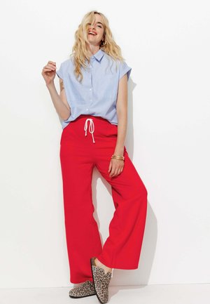 Chemise bleue à manches courtes avec des boutons, pantalons rouges à jambes larges avec un cordon blanc, et des chaussures à enfiler à imprimé léopard. Tenue simple et décontractée.