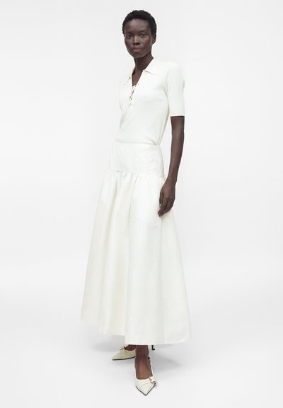 Femme portant un haut blanc côtelé à manches courtes et une longue jupe blanche taille haute avec des talons blancs à bouts pointus sur un fond uni.