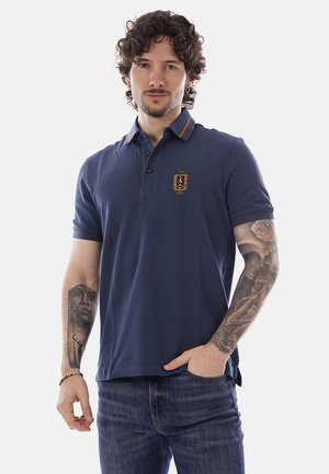 Uomo con capelli ricci scuri che indossa una polo blu navy con stemma e jeans, mostra tatuaggi su entrambi gli avambracci, mano in tasca.