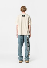 Camiseta de manga corta beige con una etiqueta negra cuadrada en la parte posterior, combinada con jeans azules que tienen una impresión gráfica lateral y detalles deshilachados.