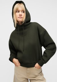 Γυναίκα με σκούρο πράσινο φούτερ με κουκούλα, με τα χέρια στις τσέπες μπεζ παντελονιού, που κοιτάζει ελαφρώς προς τα αριστερά της, μπροστά σε απλό φόντο.