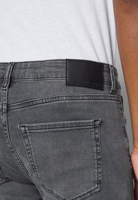Gray denim jeans with a leather back patch. Features include contrasting stitching and slim-fit design. Back pockets are visible.